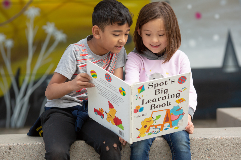 A elementary school aged boy and girl sit outside, staring at a large book.