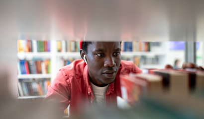 A Black college student looks for a book shelved in a campus library. He is surrounded by book stacks.