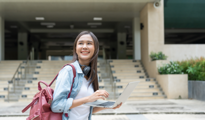 A college student holds a laptop as she stands in front of a staircase on what appears to be a college campus.