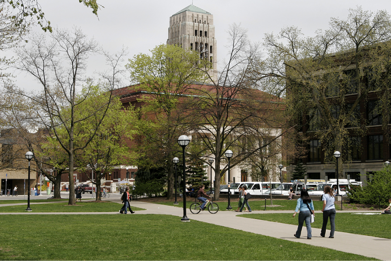 Gray skies appear over the University of Michigan campus as few walk along paved paths. They're surrounded by brick buildings and trees with sparse leaves.