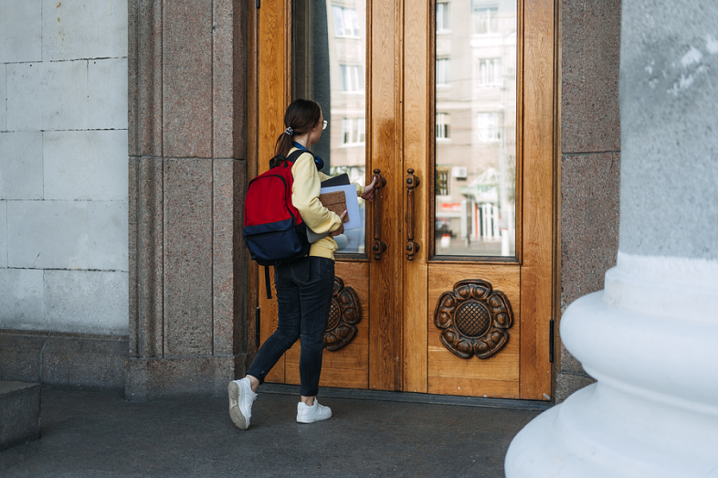 A mixed-race college students stands outside the doors of a college building. She attempts to open the double doors.