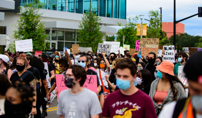 People protest outside on a busy street. They raise signs highlighting racial justices issues.