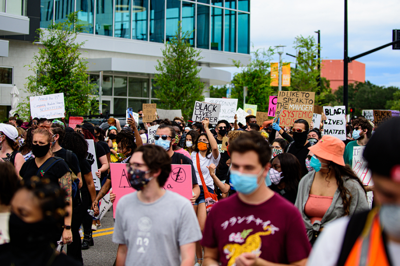 People protest outside on a busy street. They raise signs highlighting racial justices issues.