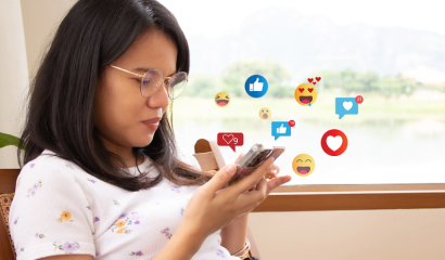 A young woman looks down at her cellphone. Social media graphics create a cloud of around her.