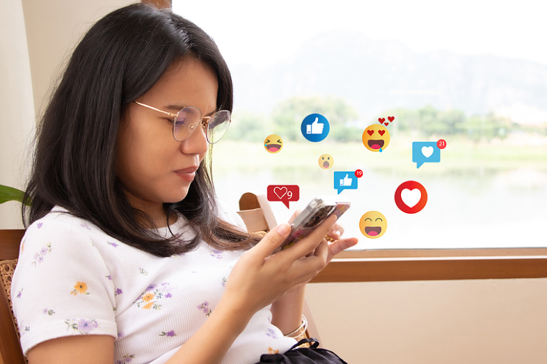 A young woman looks down at her cellphone. Social media graphics create a cloud of around her.