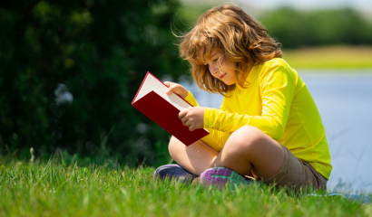 A small boy sits on green grass reading a book. A body of water is behind him.