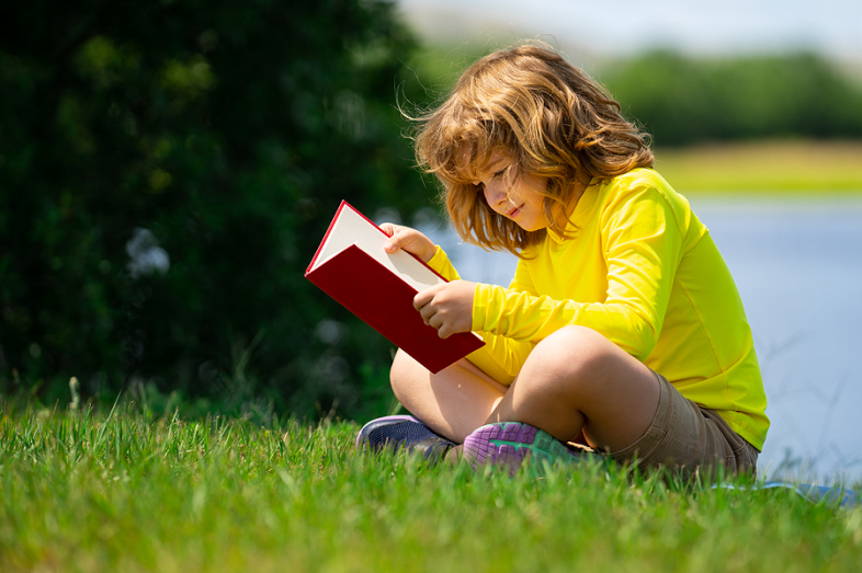 A small boy sits on green grass reading a book. A body of water is behind him.