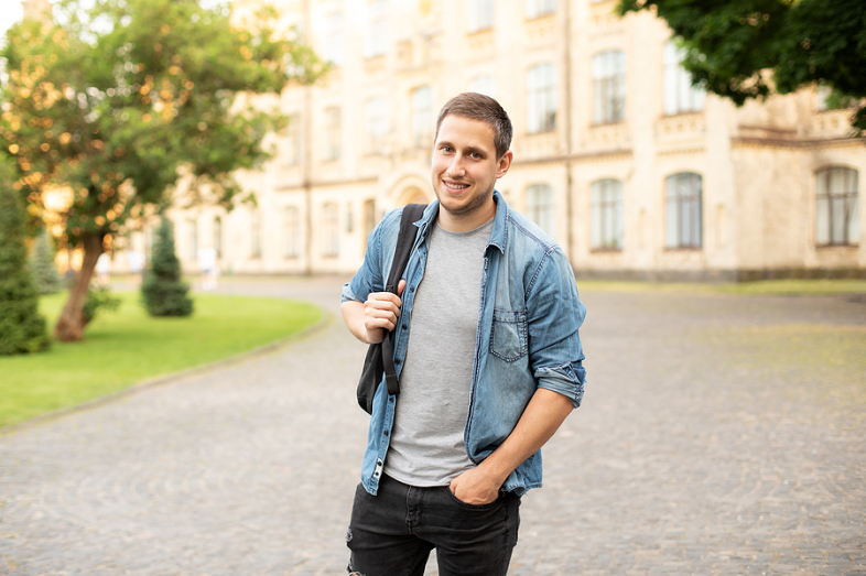 A young male stands on a college campus, with a building in the background.