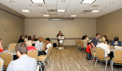 A male stands at a podium in front of a room filled with people sitting at roundtables.