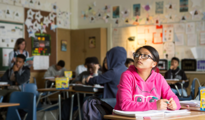 A teenage girl stares into space, pondering. She's is in a pink shirt, sitting at a desk and surrounded by classmates in her classroom.