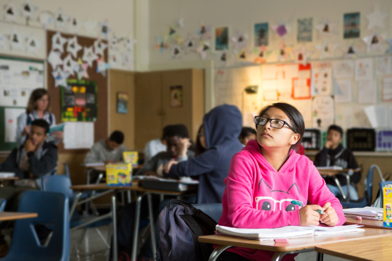 A teenage girl stares into space, pondering. She's is in a pink shirt, sitting at a desk and surrounded by classmates in her classroom.