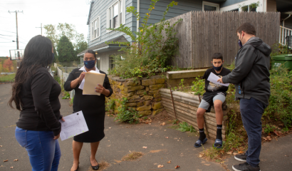 Three adults stand outside talking. A preteen sits outside his home.