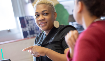 A woman points as she talks to another woman sitting next to her.